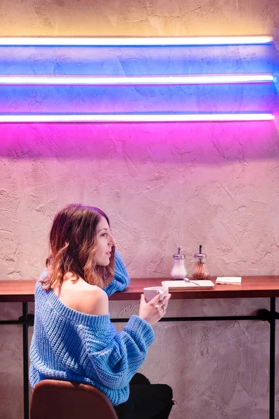 스웨터를 여자가 네온등을 가까이에 테이블에서 뜨거운 음료를 기뻐하였다 — 스톡 사진
