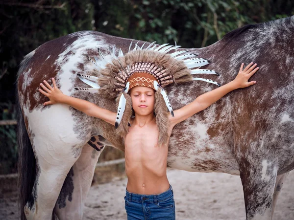 头戴印度羽毛帽 目不转睛地靠着种马 双手伸展在牧场上的有思想的孩子 — 图库照片