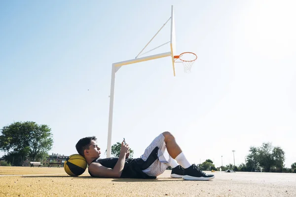 男篮球运动员在训练课后躺在黄球场上时使用智能手机 — 图库照片