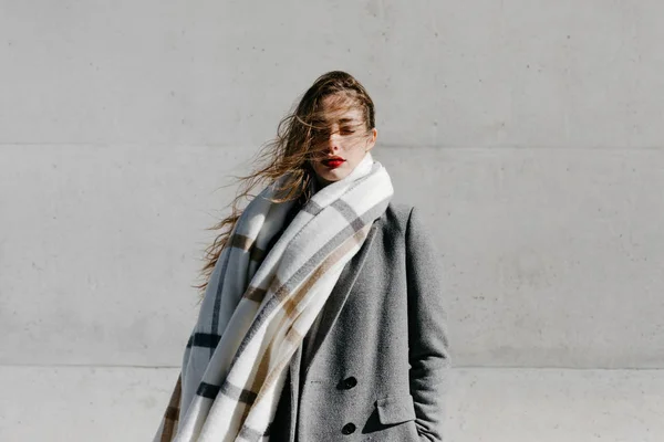 風の強い天候のコンクリートの建物の壁に対して目を閉じて立っているスタイリッシュなコートと暖かいスカーフを着た若い女性 — ストック写真