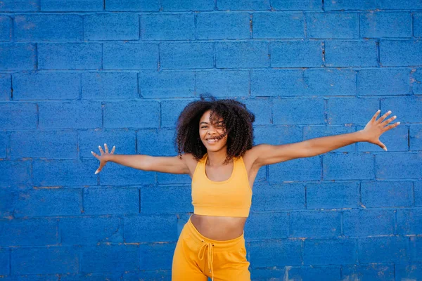 ストリートの鮮やかな青い壁を背景に腕を離れて踊る瞬間に黄色の服を着たアフリカ系アメリカ人の女性を喜ばせます — ストック写真