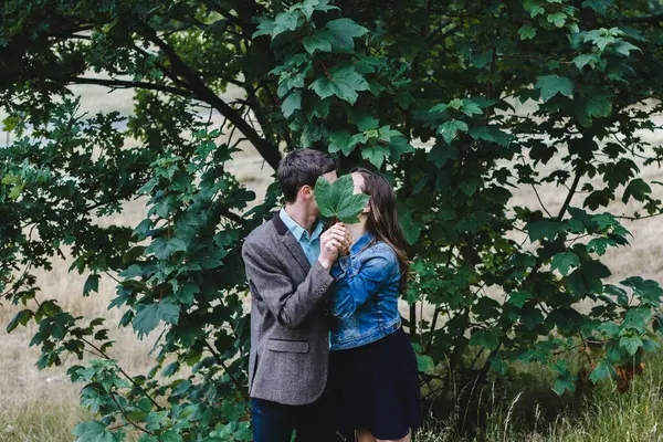 匿名のカップル抱擁とキスの側面ビューながらカバー顔とともに緑の葉で公園 — ストック写真