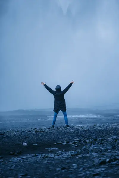 アイスランドでの休暇を楽しみながら滝を背景に腕を上げ カメラを見て立ってアウターウェアで喜びの認識できない男性観光客のバックビュー — ストック写真