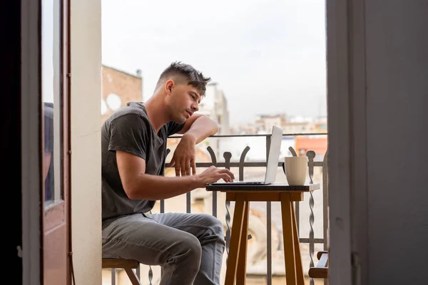 Vue latérale de l'homme adulte décontracté assis à petite table sur le balcon en utilisant un ordinateur portable et ayant une tasse de café — Photo de stock