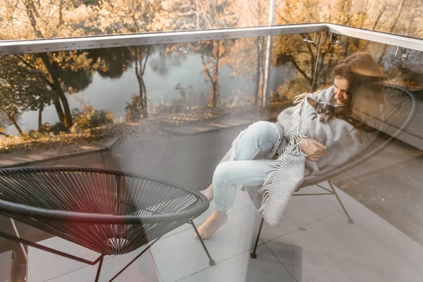 Tourné à travers le verre de la fenêtre de la femme assise sur le balcon avec le chien sur les mains au-dessus des arbres dorés de l'automne — Photo de stock