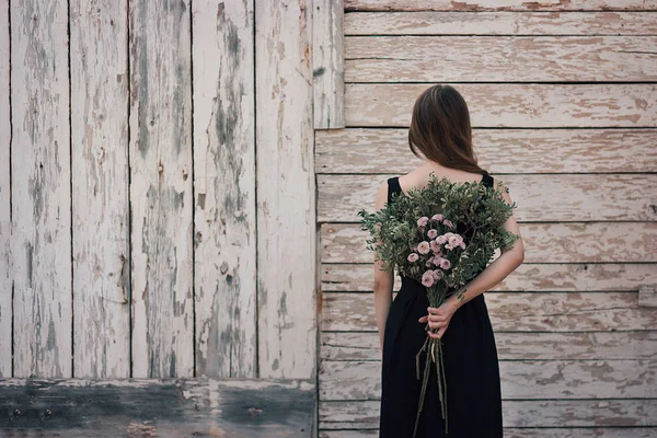 Back view of young female in elegant dress holding bunch of pretty flowers while standing near shabby wooden wall of countryside building — Stock Photo