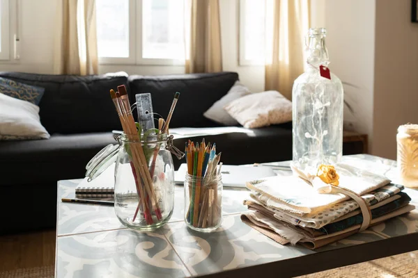 Raccolta di pennelli e matite in lattine di vetro vicino bottiglia e mucchio di materiali sul tavolo basso vicino divano in camera — Foto stock
