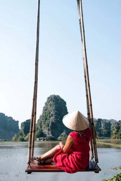 Back view of unrecognizable female tourist in casual clothes and conical hat sitting on wooden swing and admiring amazing scenery of rocks in water — Stock Photo