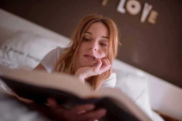 Мирна жінка відпочиває на ліжку, лежачи на животі і читаючи цікаву книжку в затишній спальні з романтичним написом на стіні і мрійливою атмосферою — стокове фото