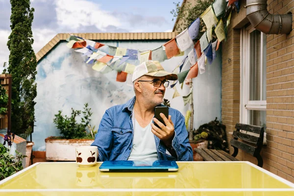 Thoughtful senior male in cap and browsing smartphone while sitting at laptop with mug of yummy hot drink in backyard of country house in sunny day — Stock Photo