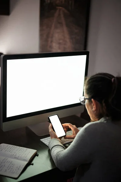 Vista lateral de una mujer joven concentrada grave en ropa casual y anteojos sentados en la mesa con computadora con pantalla blanca en blanco y teléfono inteligente de navegación mientras trabaja de forma remota por la noche en casa - foto de stock