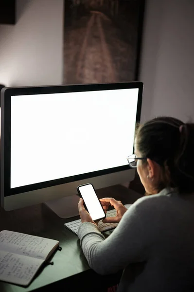 Side view of serious concentrated young female in casual wear and eyeglasses sitting at table with computer with white blank screen and browsing smartphone while working remotely at evening time at home — Stock Photo