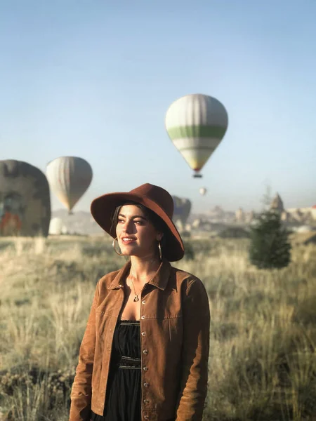 Smiling female tourist in trendy wear standing in green field and enjoying sunny weather on background of hot air balloons — Stock Photo