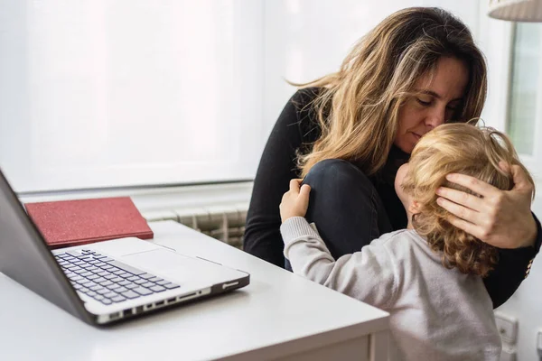 Moderna empresaria ocupada en ropa casual abrazando y besando a un niño pequeño mientras trabaja en el proyecto con el ordenador portátil en la oficina en el hogar - foto de stock