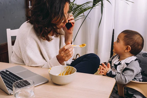 Moderna giovane lavoratrice remota che alimenta il bambino e discute di questioni commerciali sullo smartphone mentre siede a tavola con il computer portatile e lavora al progetto online — Foto stock