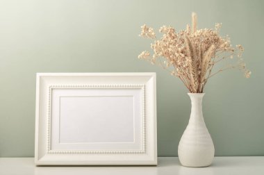 Beyaz boş fotoğraf çerçevesi ve masasında kurumuş çiçekler olan beyaz vazo. Zeytinli arka plan. Minimalist iç mekan..