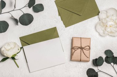 Kadınsı davetiye veya yeşil zarflı tebrik kartı modeli, hediye kutusu ve beyaz şakayık çiçekleri ve açık arkaplanda okaliptüs dalları. Boş bir kartpostal