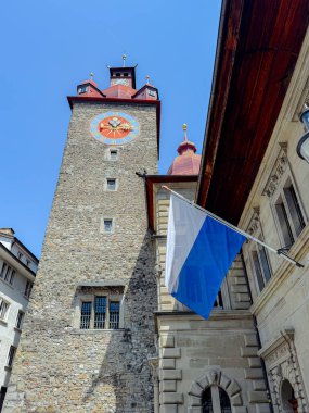 Canlı kırmızı ve altın aksanlı tarihi saat kulesi açık mavi gökyüzüne karşı dimdik ayakta duruyor. Arkasında ulusal bir bayrak dalgalanıyor ve mimariye kültürel önem katıyor..