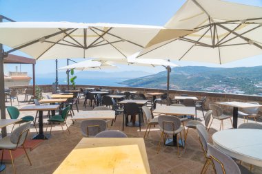 Açık kafe terasında büyük şemsiyelerin altında boş masalar ve sandalyelerle deniz ve uzak dağların çarpıcı manzarasını sunuyor. Rahatlamak ve gün ışığında yemek yemek için ideal ortam..