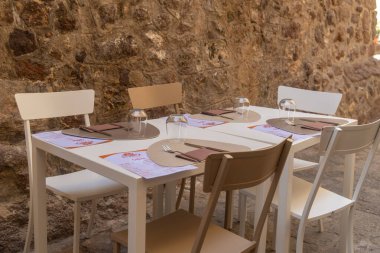 Açık hava yemek masası seti. Dört sandalye, taş bir duvara karşı düzgünce dizilmiş. Her yer menüler, peçeteler ve bardaklarla dolu.