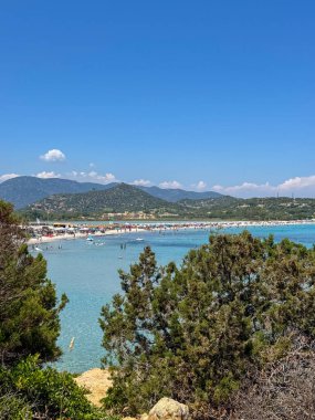 Sardinya 'da popüler bir plajda güneşli bir yaz günü. Turkuaz sular, güneş banyosu yapanlar ve uzak yeşil dağlar..