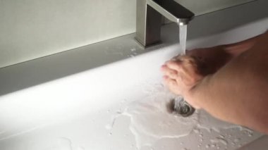 Genç bir adam ellerini yıkar. Beyaz lavabo ve paslanmaz musluk..