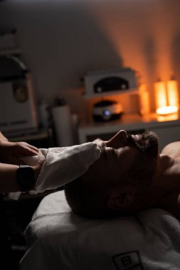 Bir adam masaj masasında havluyla kaplıyken yüz spa tedavisi görüyor. Bir profesyonel nazikçe yüzüne havlu yapıştırır ya da sıkıştırır. Atmosfer sakin ve rahatlatıcı, sıcak, yumuşak ışıklandırmayla.