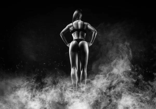 Image of a female athlete in a fire arena surrounded by smoke. Back view. Bodybuilding and fitness concept. Mixed media
