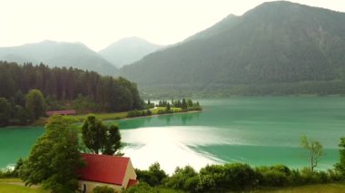 Turkuaz alp gölü, orman kıyısı ve bulutlu gökyüzünün altında kırmızı çatı manzarası.