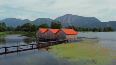 Ahşap kayıkhaneler, kırmızı çatılar ve dağlarla göl manzarası.