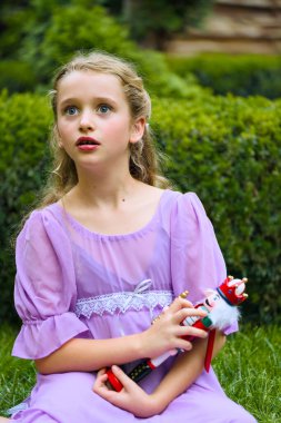 Mor elbiseli bir çocuk elinde fındıkkıran bebeğiyle oturmuş, rüya gibi bir ifadeyle yukarı bakıyor. Sihirli Noel tatili konsepti, çocukluk hayalgücü ve açık havada peri masalı anı..