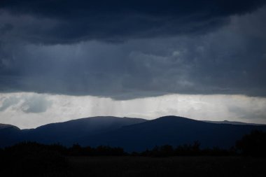 Kara fırtına bulutları siluetli dağların tepelerinde toplanırken, dar bir ışık bandı gökyüzünden düşen bir yağmur perdesini gözler önüne seriyor. Gölge ve ışık arasındaki dramatik zıtlık sahneye güçlü bir sinematik kalite katar..