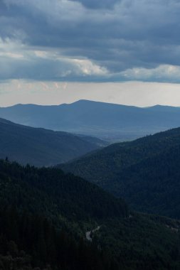 Yoğun ormanlarla kaplı yuvarlanan dağ sırtları, dramatik fırtınalı bir gökyüzü tepemizde belirdikçe, gözü uzak bir vadiye ve kasabaya yönlendirir. Aşağı ormanı kesen dolambaçlı bir yol bu uçsuz bucaksız doğal araziye derinlik ve ölçek ekliyor..