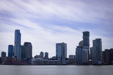 New York 'un Aşağı Manhattan siluetinin limanın karşısından çekilmiş panoramik görüntüsü. Görüntüde, en tanınmış finans binalarından bazıları da dahil olmak üzere mavi gökyüzü altındaki modern ve tarihi gökdelenlerin bir karışımı yer alıyor