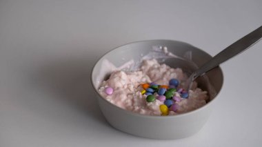 Beyaz seramik kâse süzme peynirli tatlı, renkli çikolata şekerleri ve tarafsız bir zeminde kaşık. Sağlıklı kahvaltı, çocuk beslenme, gıda tasarımı, süt ürünleri veya yaratıcı atıştırmalık kimliği kavramlarını resmetmek için idealdir.