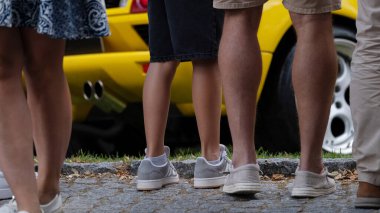 Sarı bir spor arabanın yakınındaki kaldırımda duran insanların bacak ve ayakkabılarının görüntüsünü kapatın. Görüntü, yaşam tarzı, otomotiv kültürü, turizm, etkinlikler, seyirciler, şehir hayatı ve sosyal toplanma konularında kullanılabilir..