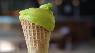 Koyu yeşil bir waffle külahında açık yeşil şam fıstıklı dondurma, sıcak iç ışıkları yansıtan cam camlı modern bir yapının önünde tutularak hafifçe eriyor. Yüksek kalite 4k