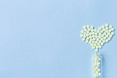 Sarı yuvarlak tabletler, mavi bir zemin üzerinde cam bir şişeden dökülen kalp şeklinde yatar. Sağlık, tedavi ve hastalıkların önlenmesi kavramı. Mesaj için yer var. Seçici odak