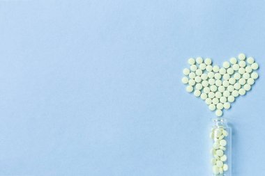 Sarı yuvarlak tabletler, mavi bir zemin üzerinde cam bir şişeden dökülen kalp şeklinde yatar. Sağlık, tedavi ve hastalıkların önlenmesi kavramı. Mesaj için yer var. Seçici odak