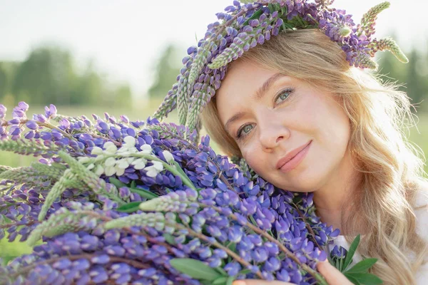 Çiçekli bir çelenk içindeki sarışın bir kız lupine buketi ile nazikçe gülümser ve güneşli bir yaz gününde çiçek çayırındaki kameraya bakar, çiçeklerin kokusunu içine çeker.
