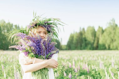 Çiçek çelenginde mutlu bir kız bir buket yabani çiçeği kucaklıyor ve gülümsüyor, çiçek kokusunu içine çekiyor, bir buket lupin. Genç bir kızın neşeli görünüşü