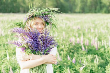 Çiçek çelenginde mutlu bir kız bir buket yabani çiçeği kucaklıyor ve gülümsüyor, çiçek kokusunu içine çekiyor, bir buket lupin. Genç bir kızın neşeli görünüşü