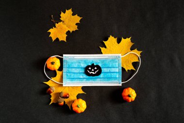 Salgın sırasında Cadılar Bayramı. Uzayı kopyala Yukarıdan manzara. 2020 sonbaharında. Siyah kağıttan siluetler, yarasa, balkabağı, örümcek içeren siyah arka planda tıbbi bir maskeyle sarı akçaağaç yaprakları.