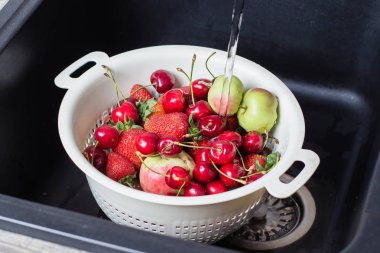 colander with ripe cherries and apples with strawberries in black kitchen-sink under stream water, Summer food concept clipart