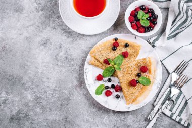 Lezzetli krepler kahvaltı gri beton tablo arka plan üzerinde. Ortodoks tatil Maslenitsa. Krep ile dut Siyah frenk üzümü, ahududu, kavanoz bal ve nane. Beyaz fincan da çay. Üstten Görünüm, kopya alanı