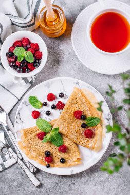 Lezzetli krepler kahvaltı gri beton tablo arka plan üzerinde. Ortodoks tatil Maslenitsa. Krep ile dut Siyah frenk üzümü, ahududu, kavanoz bal ve nane. Beyaz fincan da çay. Üstten Görünüm