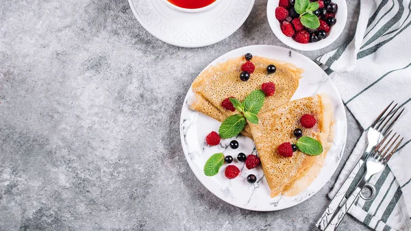 Delicious Crepes Breakfast on gray concrete table background. Orthodox holiday Maslenitsa. Pancakes with berry black currant, raspberry, jar of honey and mint. White cup of tea. Top view, copy space,