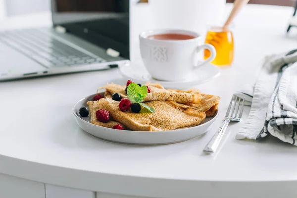 Delicioso desayuno. Mesa de escritorio blanca con portátil y crepes frescos panqueques, taza de té y tarro de miel. Concepto freelancer. Comida saludable para el desayuno . — Foto de Stock