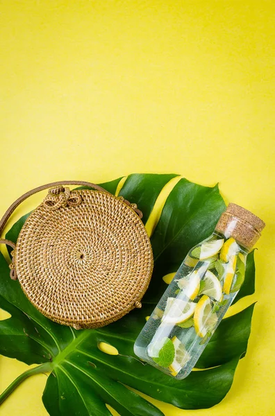 Round rattan bag and glass bottle with lemonade on a Monstera leaf. Yellow, sunny background. Flat lay, top view