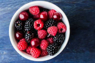 Mavi ahşap masa arka plan üzerinde Yaz taze çilek ile beyaz fincan üst görünümü. Kopya alanı. Vejetaryen gıda - ahududu, böğürtlen ve kiraz. Rustik stil